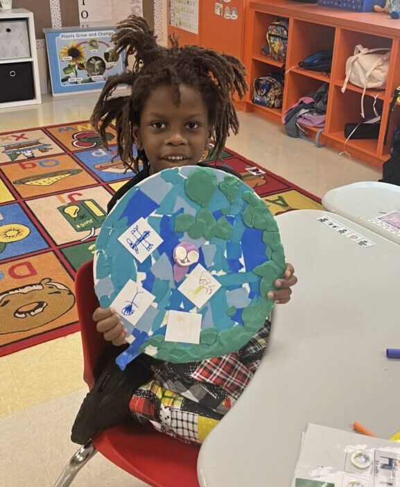 A young girl holding a paper globe in front of a classroom at the Center for Creative Education.