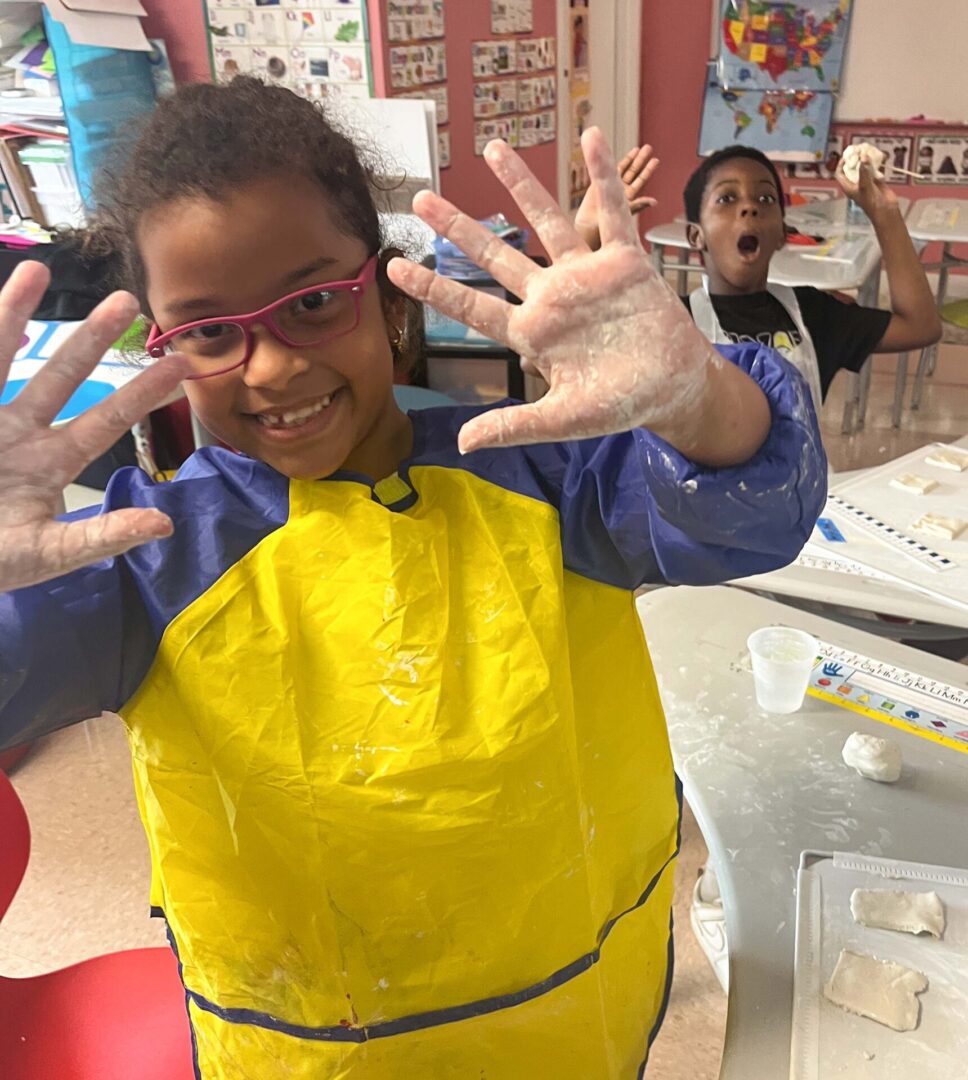 A girl in a yellow apron at the center for creative education with her hands up in the air.