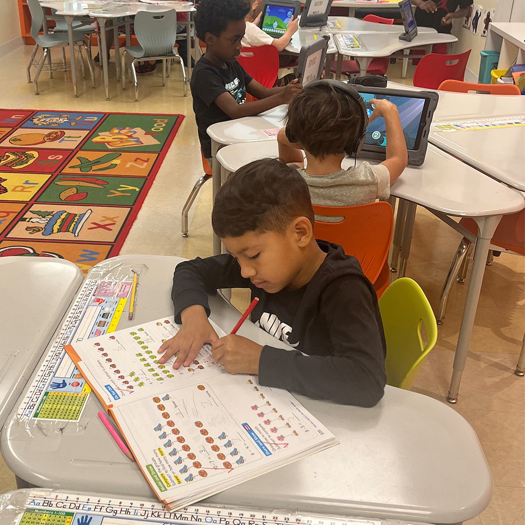 Children in a playground setting; one focused on writing in a workbook at a desk while others use computers in the background.