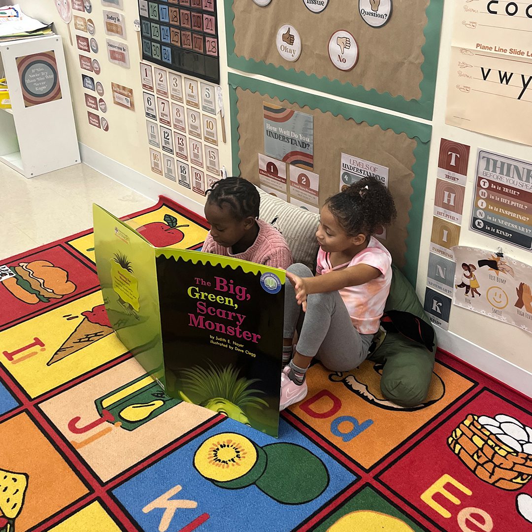 Two young children reading a large popup book called "The Big Greig, Sean, Monster" on a colorful educational rug in a playground.