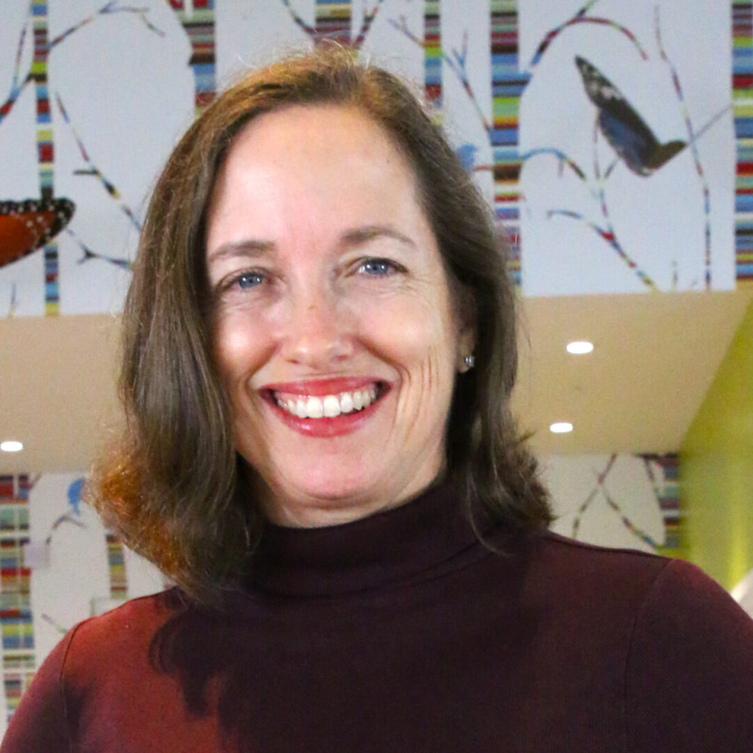 A woman, who is one of the teaching artists, wears a burgundy turtleneck as she smiles in front of a wallpapered room.