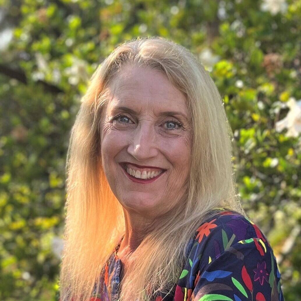 A woman with long blonde hair smiling in front of trees, teaching artists.