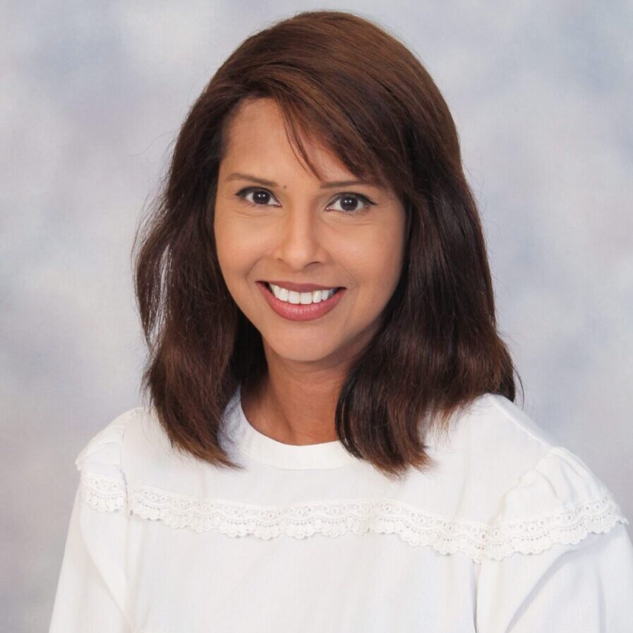 Portrait of a smiling woman with shoulder-length brown hair, wearing a white blouse and holding a staff, against a mottled blue and gray background.