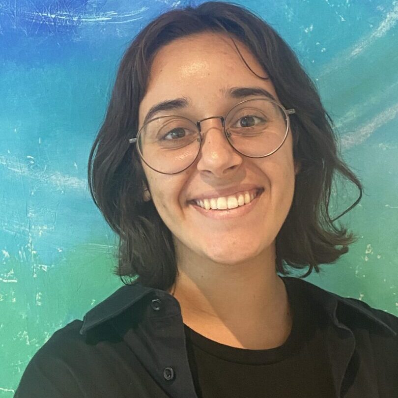 A woman wearing glasses smiles in front of a colorful wall, capturing the vibrant essence of teaching artists.