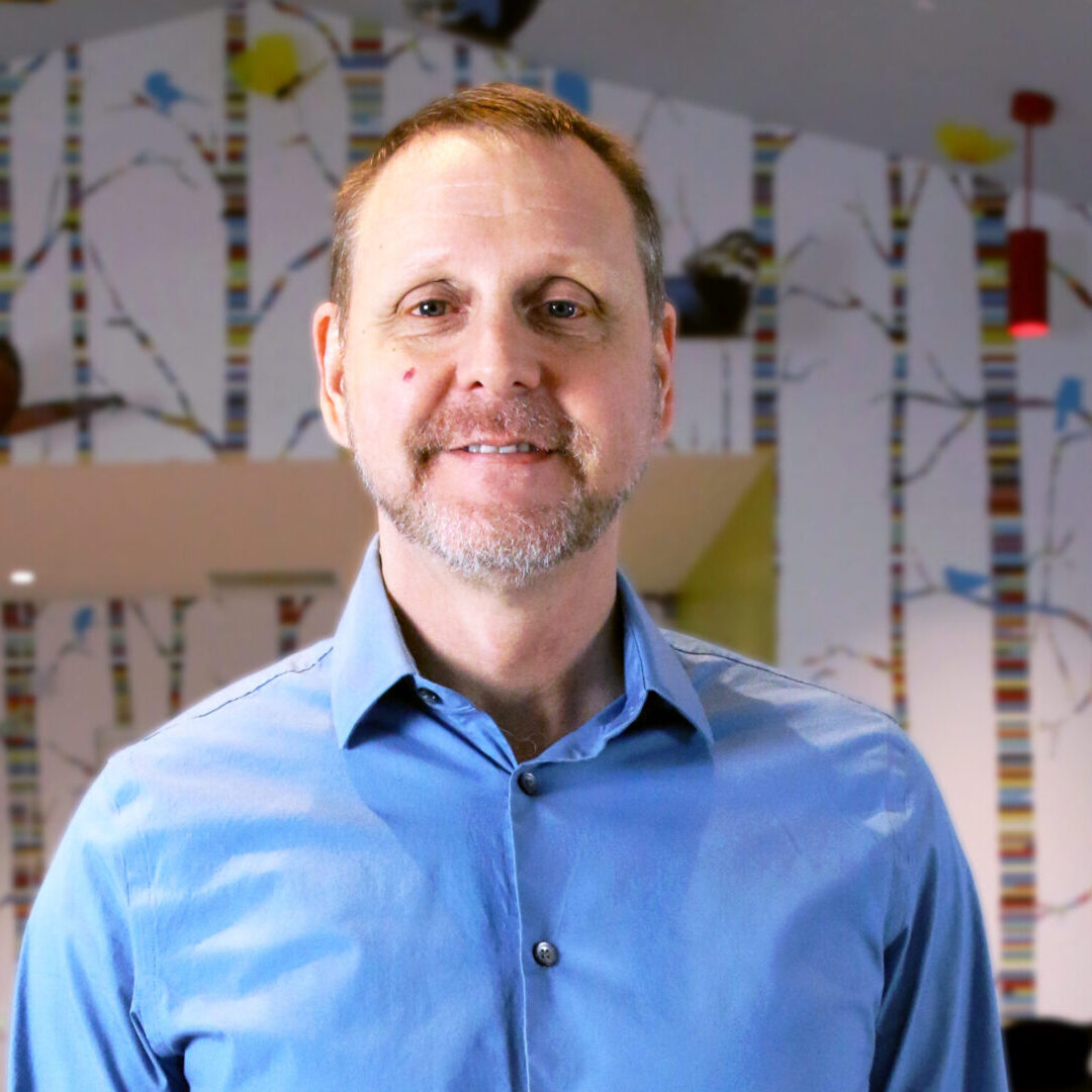 A staff member in a blue shirt standing in front of a wallpapered room.