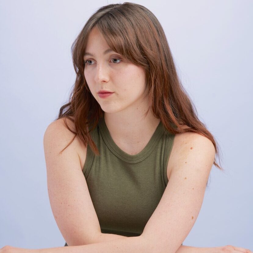 A young woman with long brown hair, wearing a green tank top and identified as one of the teaching artists, sits at a table looking to her right with a thoughtful expression.