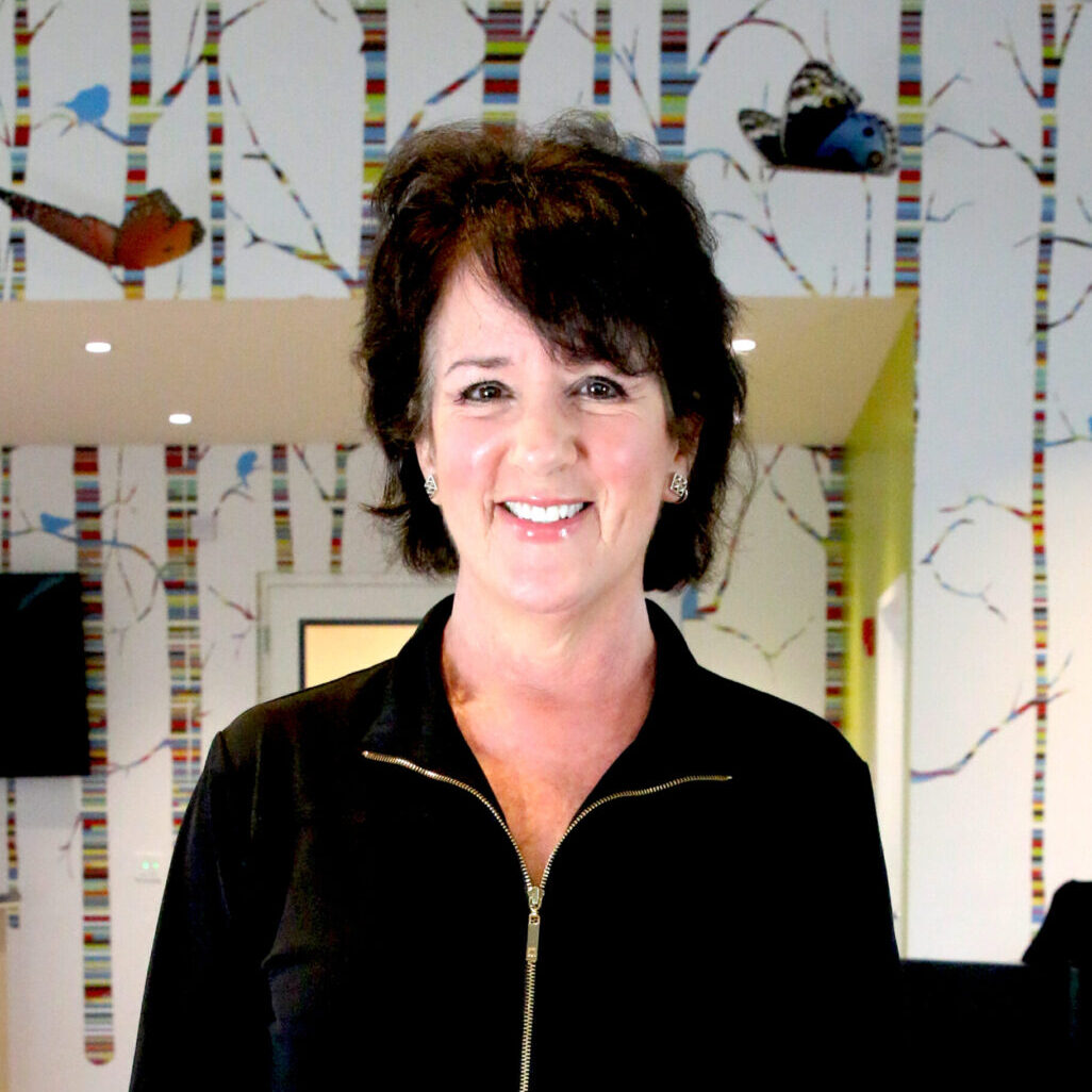 A teaching artist in a black jacket standing in front of a wallpapered room.