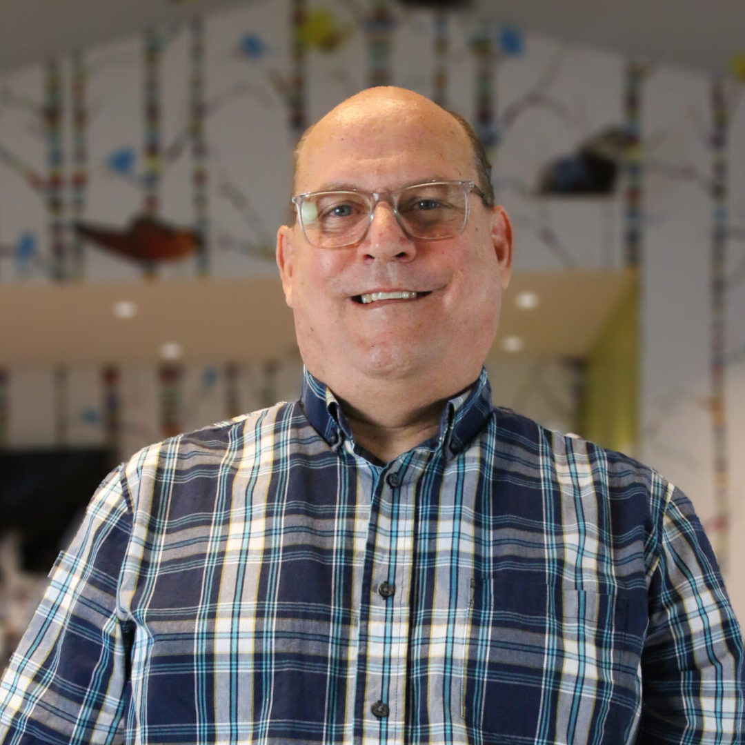 A bald man in a plaid shirt, one of the teaching artists, standing in front of a wall.