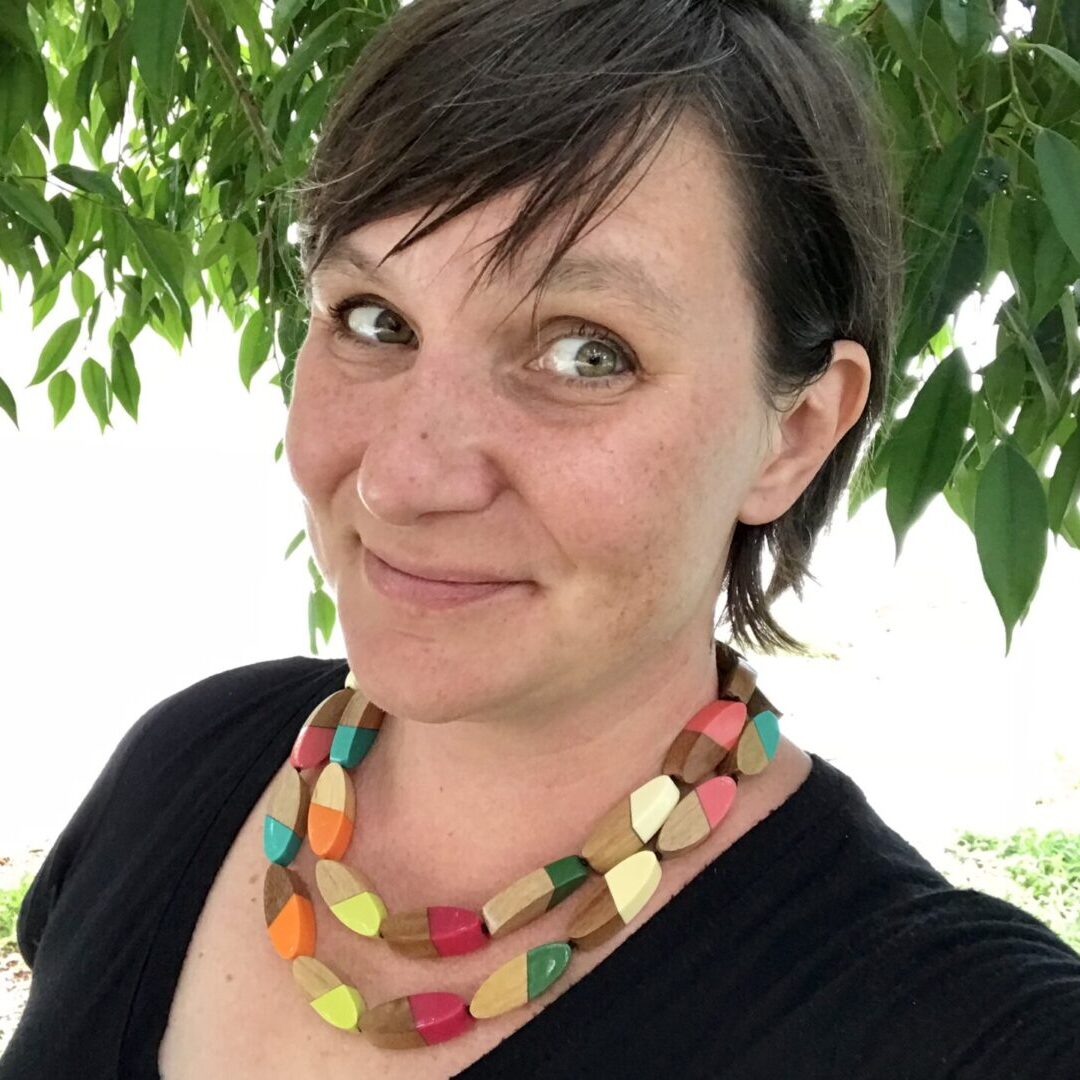 A woman, who is one of the teaching artists, wearing a colorful necklace in front of a tree.