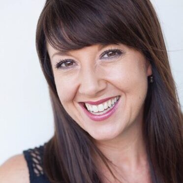 A teaching artist with long brown hair smiling in front of a white wall.