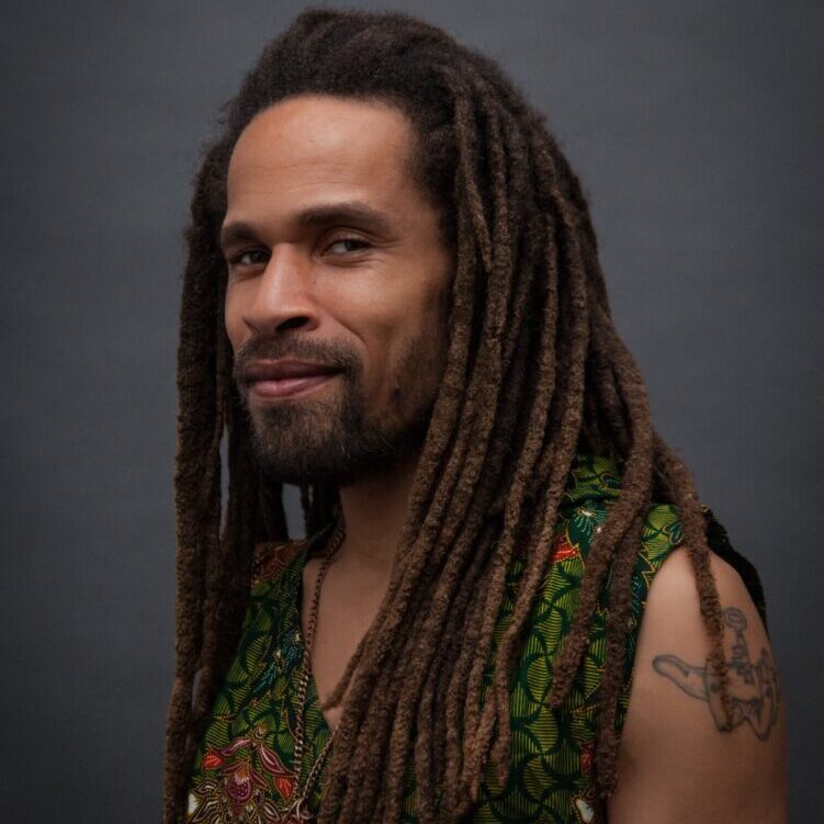 A teaching artist with long dreadlocks wearing a green patterned shirt and a subtle smile, posing against a gray background.